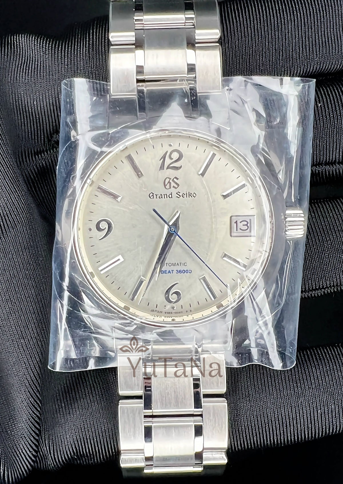 A Grand Seiko stainless steel wristwatch with a silver dial and metal bracelet, still sealed in clear protective plastic wrap.