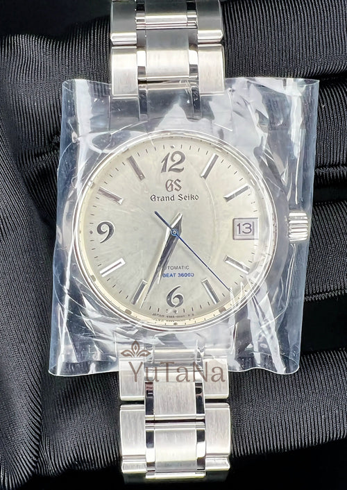 A Grand Seiko stainless steel wristwatch with a silver dial and metal bracelet, still sealed in clear protective plastic wrap.