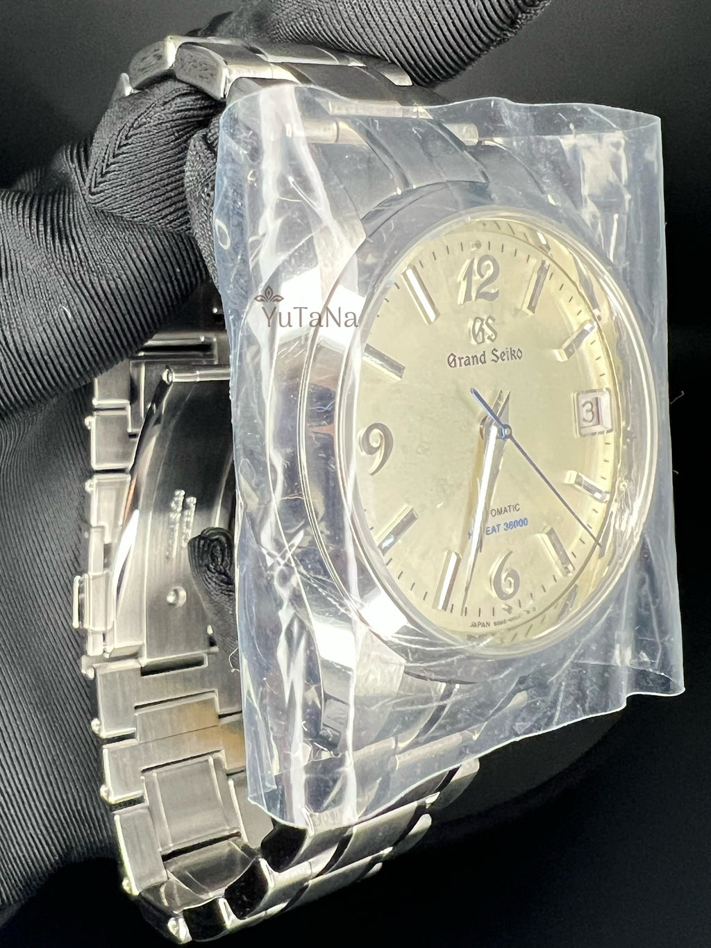 A Grand Seiko stainless steel wristwatch with a cream-colored dial, held by a black-gloved hand inside a clear protective plastic bag. The watch features a silver-tone metal link bracelet and a date window at the 3 o'clock position.