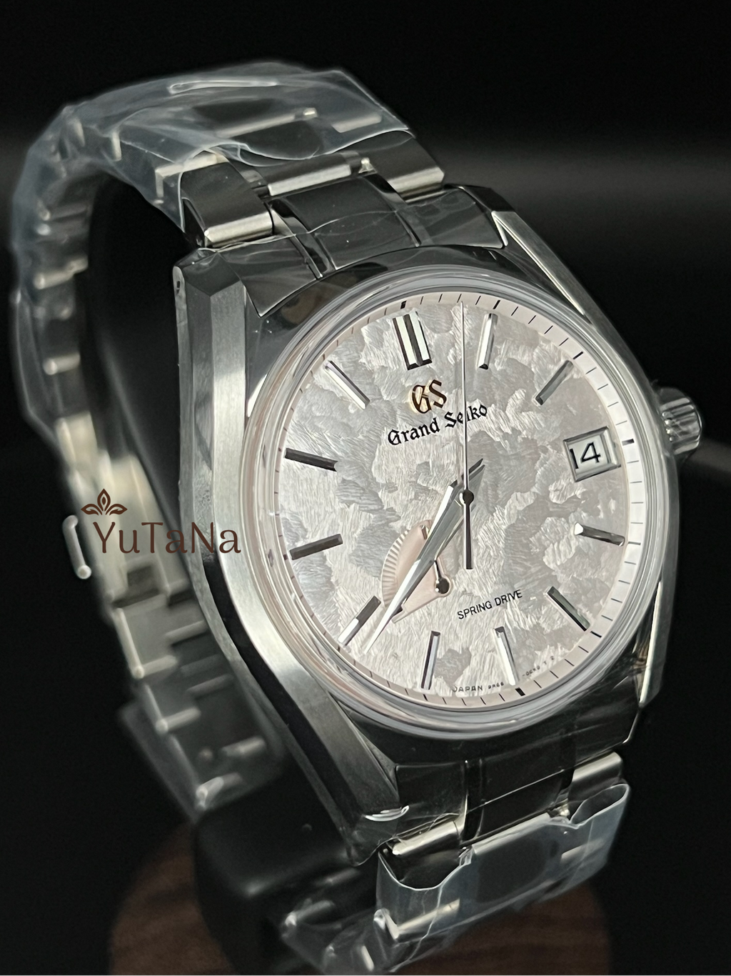 A Grand Seiko wristwatch with a silver-toned metal bracelet and case, featuring a textured silver dial with a date window and Roman numeral hour markers, shown on a dark background.