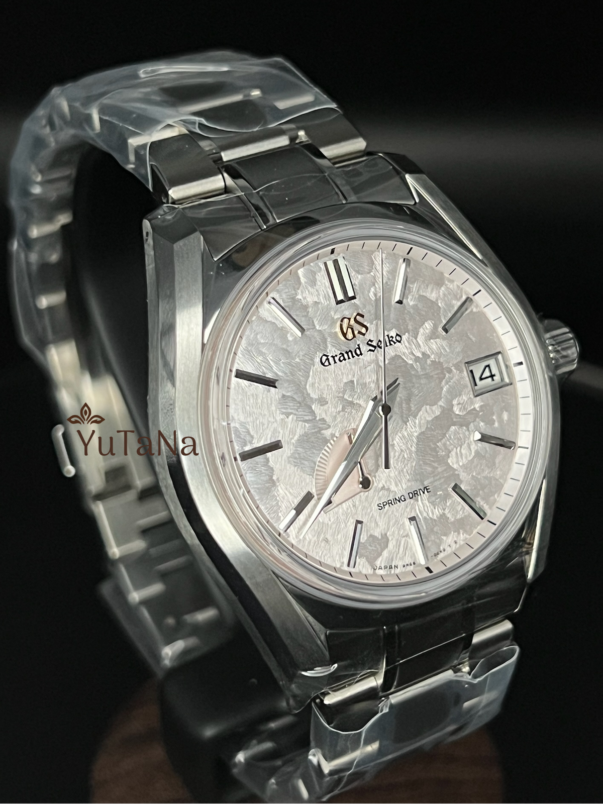 A Grand Seiko wristwatch with a silver-toned metal bracelet and case, featuring a textured silver dial with a date window and Roman numeral hour markers, shown on a dark background.