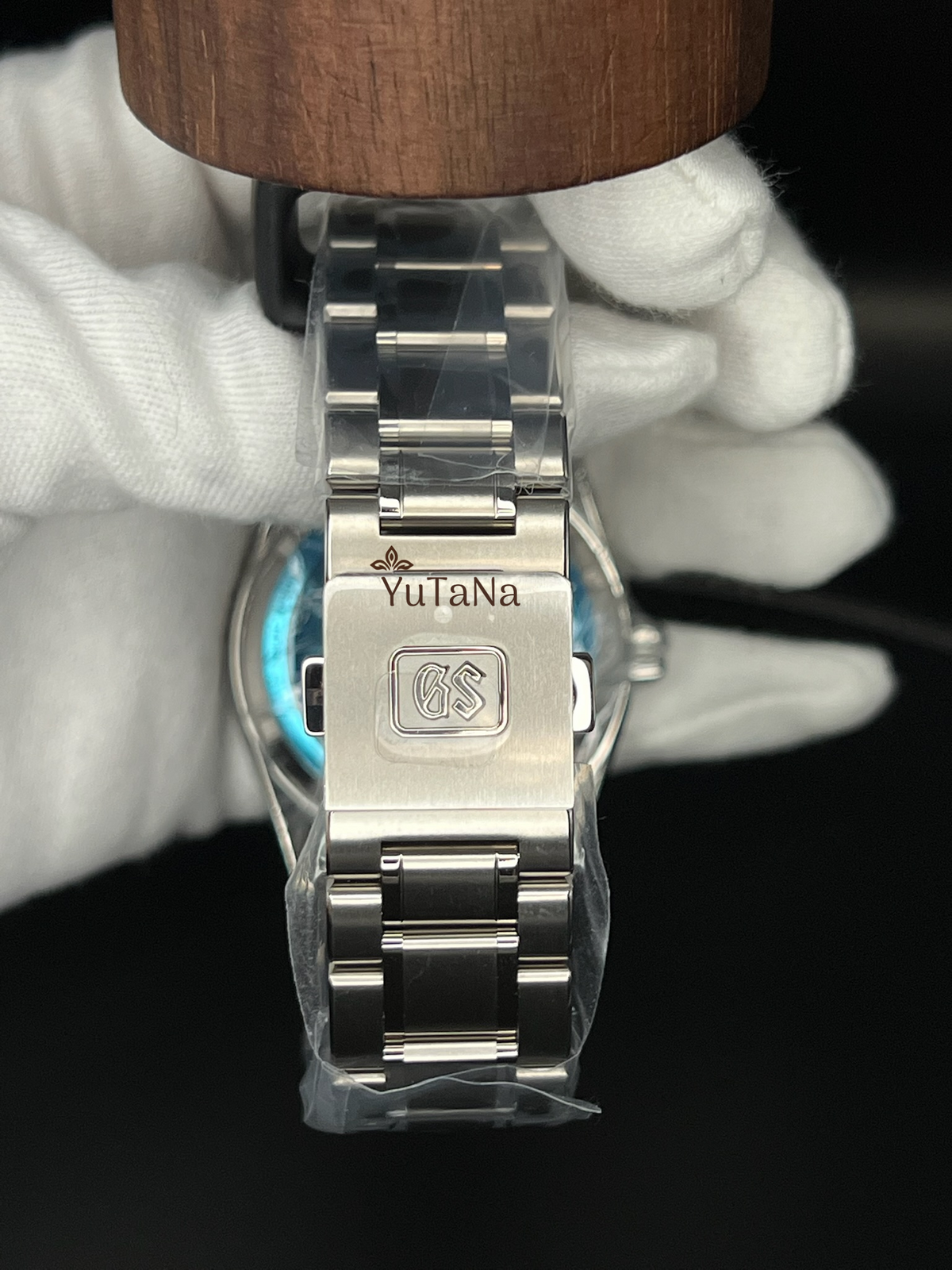 Close-up of the back of a stainless steel watch with a metal bracelet, held by white-gloved hands against a black background. The watch case and bracelet are silver-toned.