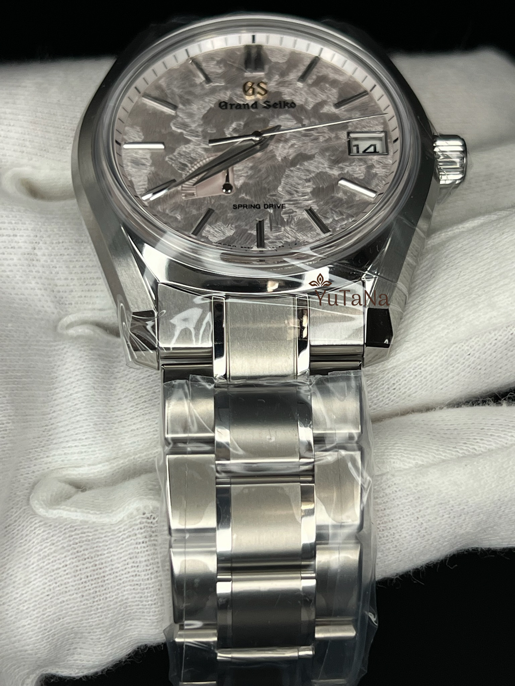 A Grand Seiko wristwatch with a silver stainless steel case and link bracelet, featuring a textured gray dial with a date window and a white-gloved hand holding it.