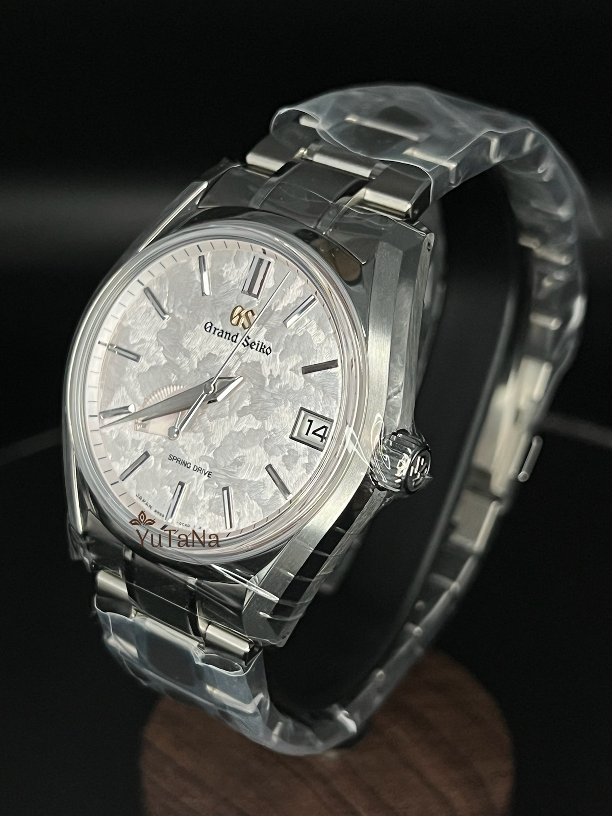 A Grand Seiko Shunbun wristwatch with a silver-toned stainless steel case and bracelet, featuring a textured white dial with gray marbled details, a date window, and protective plastic wrapping on the bracelet, displayed on a dark background.