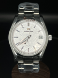 A Grand Seiko wristwatch with a silver stainless steel case and bracelet, featuring a textured white dial with Roman numerals, a date window, and protective plastic wrapping on the links, set against a dark background.
