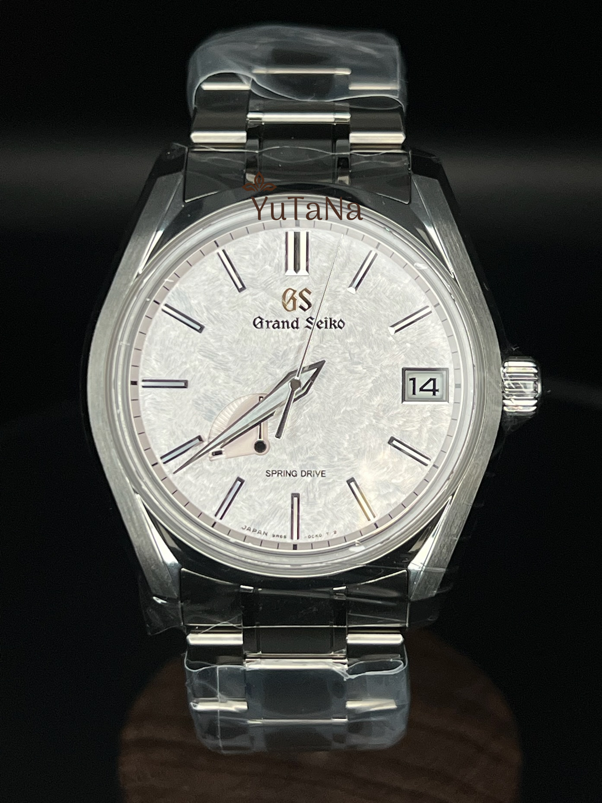 A Grand Seiko wristwatch with a silver stainless steel case and bracelet, featuring a textured white dial with Roman numerals, a date window, and protective plastic wrapping on the links, set against a dark background.