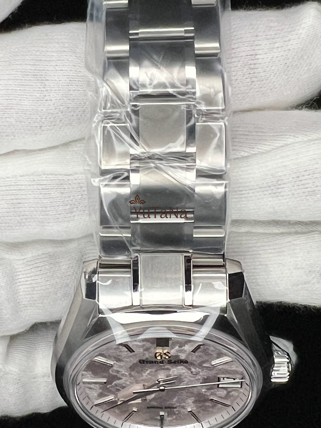 A close-up of a stainless steel wristwatch with a silver-toned metal bracelet, wrapped in protective plastic. 