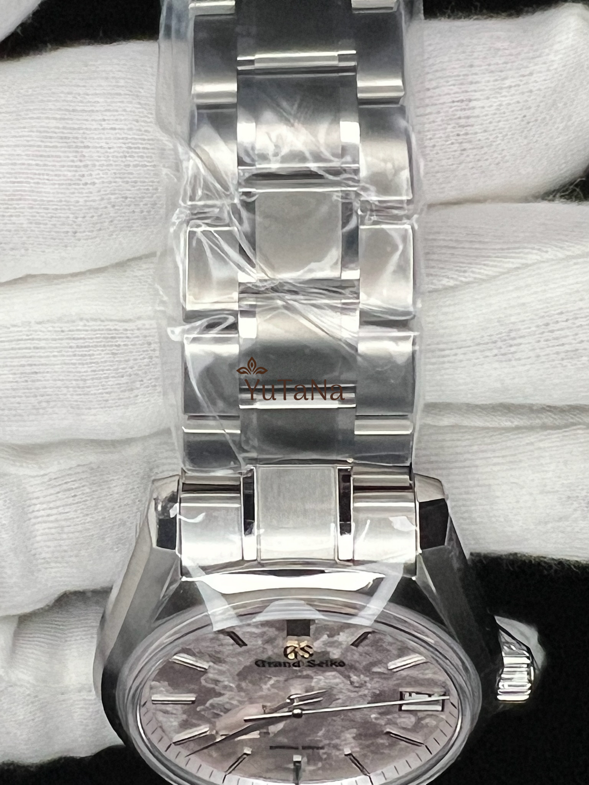 A close-up of a stainless steel wristwatch with a silver-toned metal bracelet, wrapped in protective plastic. 