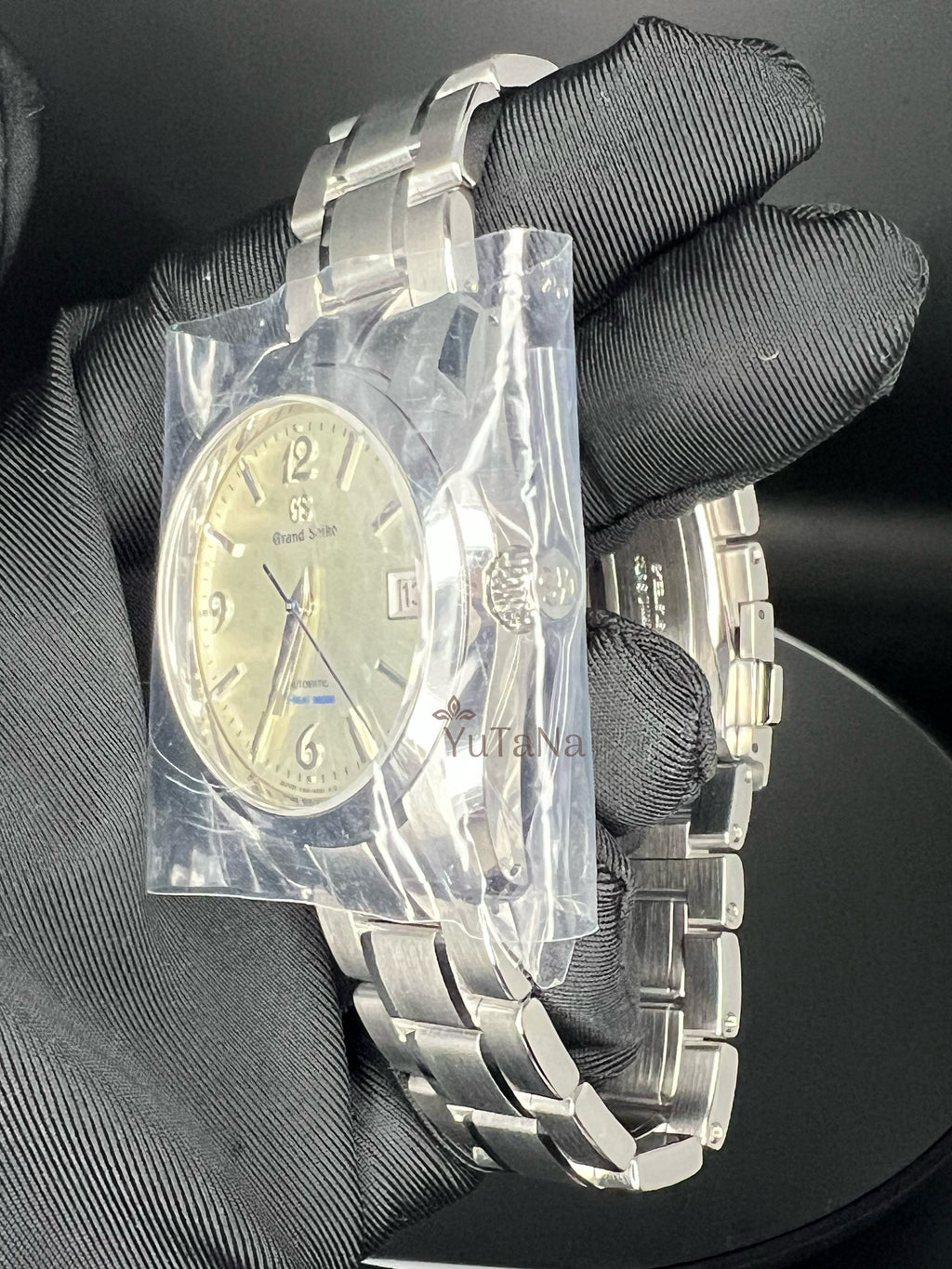 A stainless steel wristwatch with a silver-tone metal bracelet and a champagne-gold dial, held by a hand wearing a black textured glove. The watch is still sealed in a protective plastic bag wrapped around the watch case.