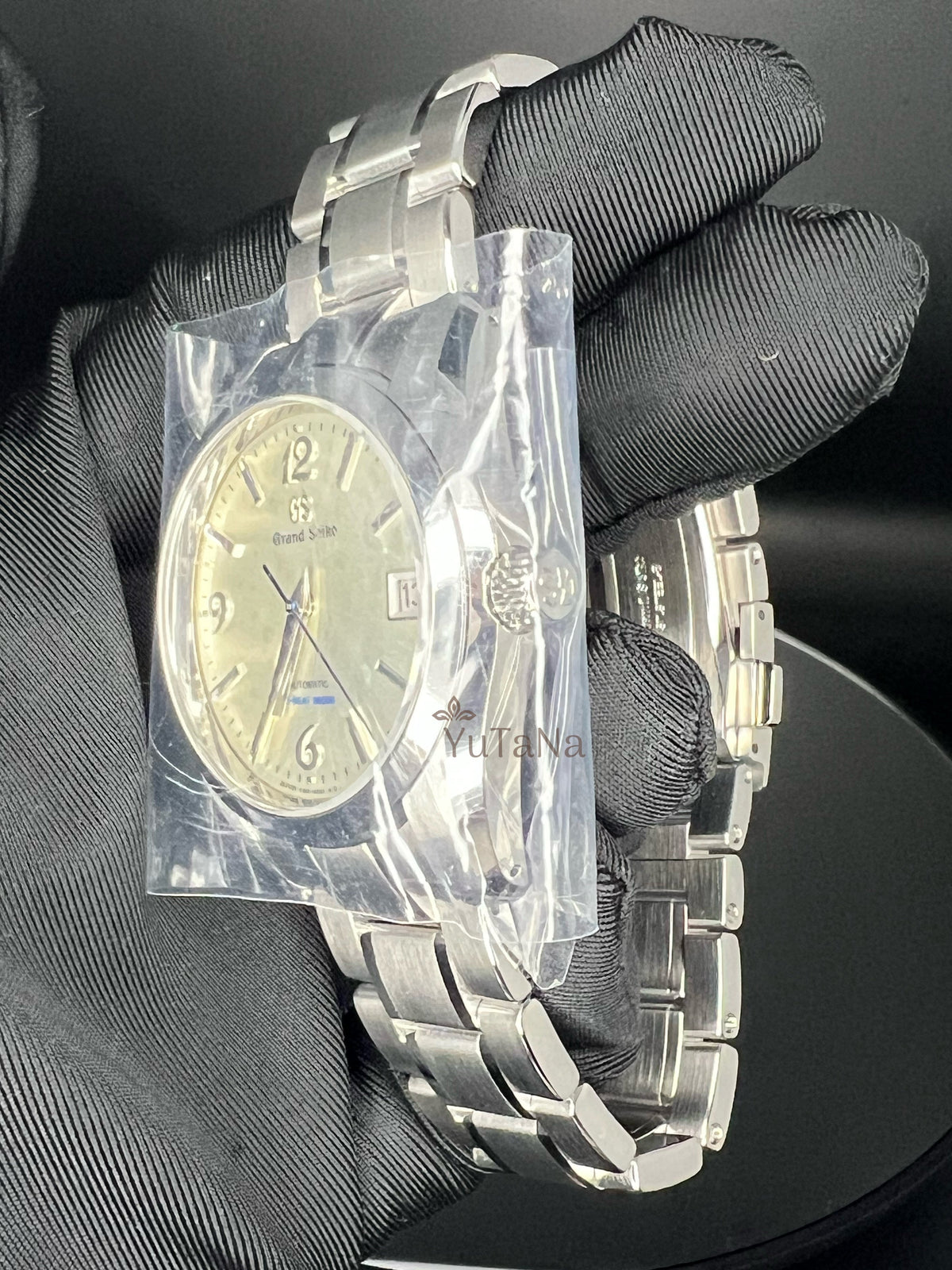 A stainless steel wristwatch with a silver-tone metal bracelet and a champagne-gold dial, held by a hand wearing a black textured glove. The watch is still sealed in a protective plastic bag wrapped around the watch case.